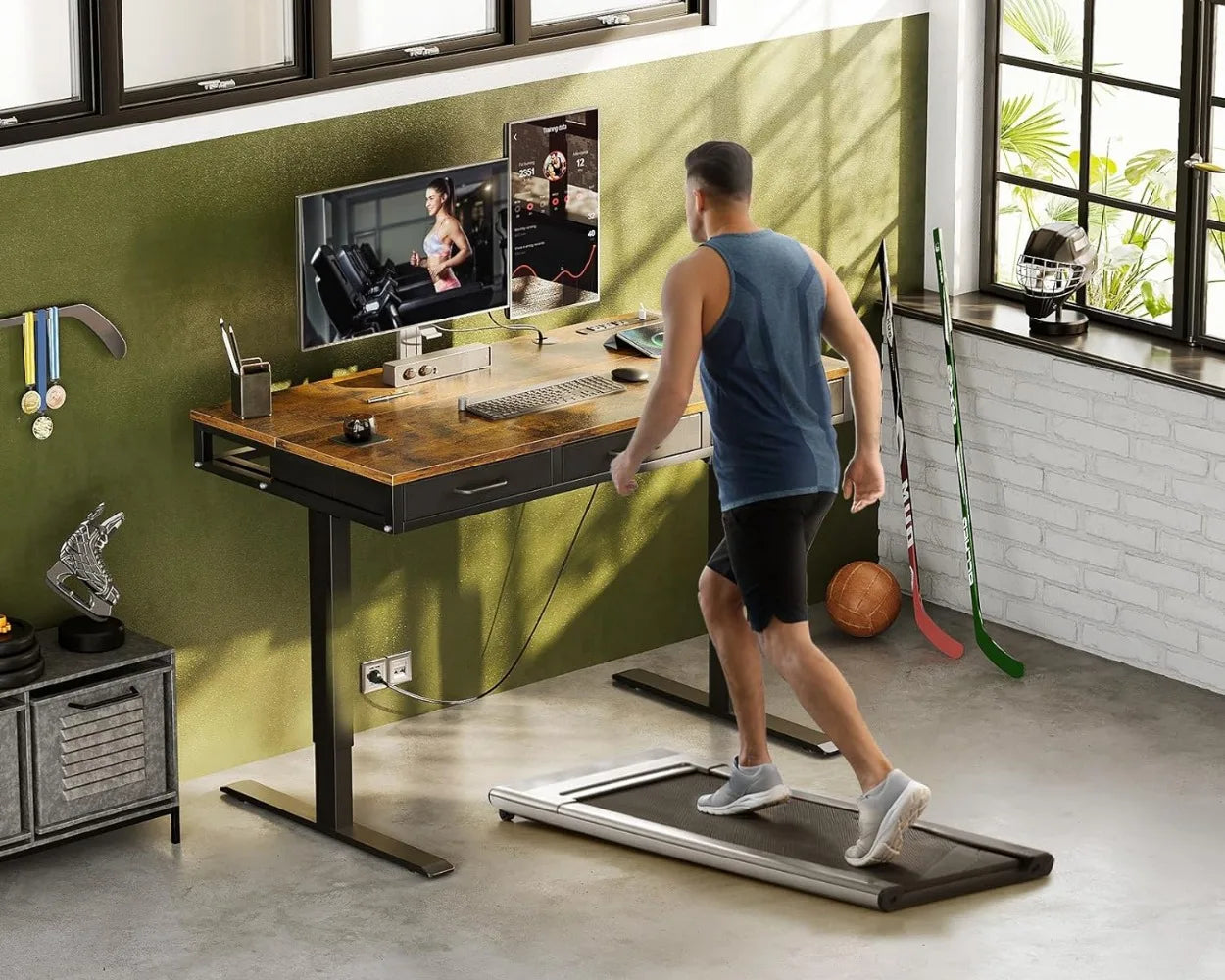 Electric standing desk with oak finish in a home office gym setup, featuring dual monitors, storage drawers, and a treadmill for an active, ergonomic workspace.